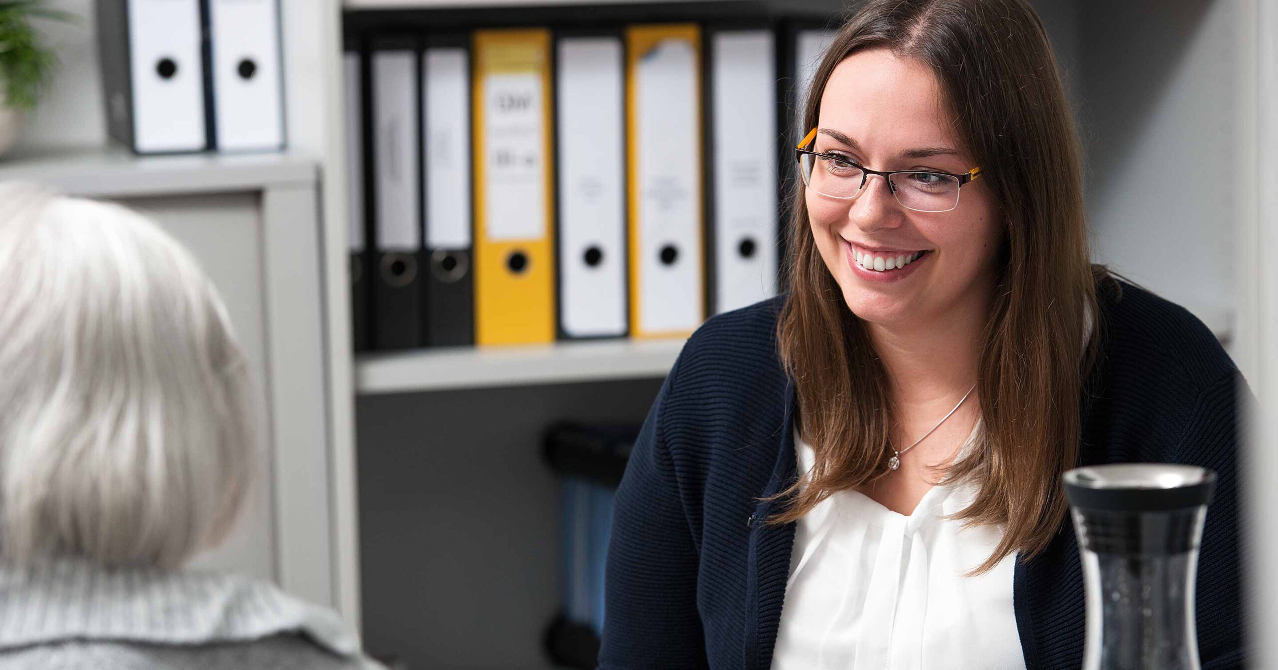 Eine Frau mit Brille, weißer Bluse und dunkler Strickjacke sitzt lächelnd an einem Schreibtisch. Sie unterhält sich mit einer anderen Person, die mit dem Rücken zur Kamera steht. Im Hintergrund sind Büroregale mit Ordnern zu sehen.