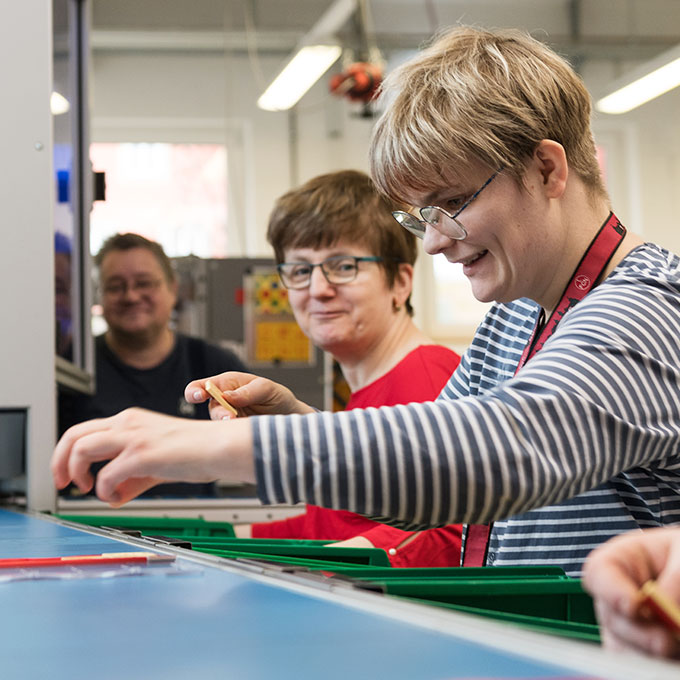 Eine Gruppe von vier Personen lächelt und arbeitet an einem Tisch mit grünen Tabletts. Eine Person im gestreiften Hemd steht im Vordergrund und konzentriert sich auf ihre Aufgabe. Der Raum ist hell beleuchtet und im Hintergrund sind Maschinen zu sehen.
