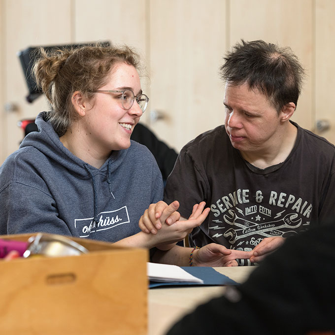 Eine junge Frau mit Brille und Kapuzenpullover lächelt und hält Händchen mit einem Mann in einem grauen „Service & Repair“-T-Shirt. Sie sitzen an einem Tisch und unterhalten sich.