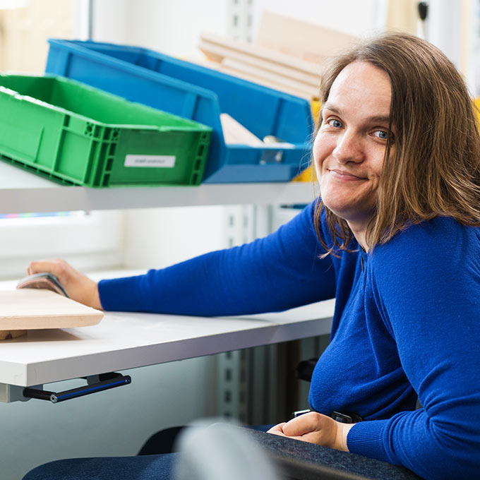 Eine Frau im blauen Pullover sitzt an einem Schreibtisch und lächelt in die Kamera. Sie befindet sich in einem hellen Raum mit Regalen und bunten Behältern um sie herum.