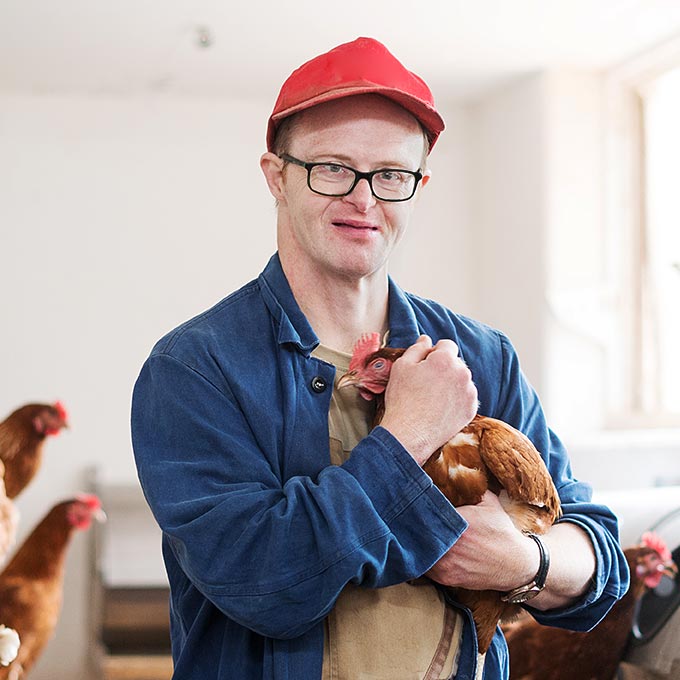 Mann in blauer Jacke und roter Mütze lächelt und hält ein braunes Huhn in einem Raum mit weiteren Hühnern im Hintergrund.