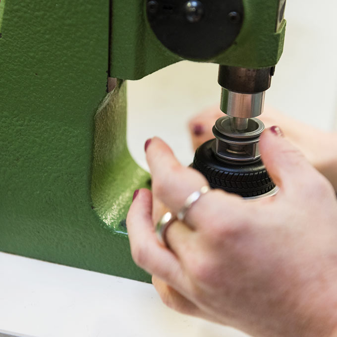 Nahaufnahme von Händen, die eine grüne Industriemaschine bedienen und ein Metallteil pressen. Die Finger der Person liegen auf einem schwarzen, geriffelten runden Stück. Die Maschine ist auf einer weißen Oberfläche montiert.