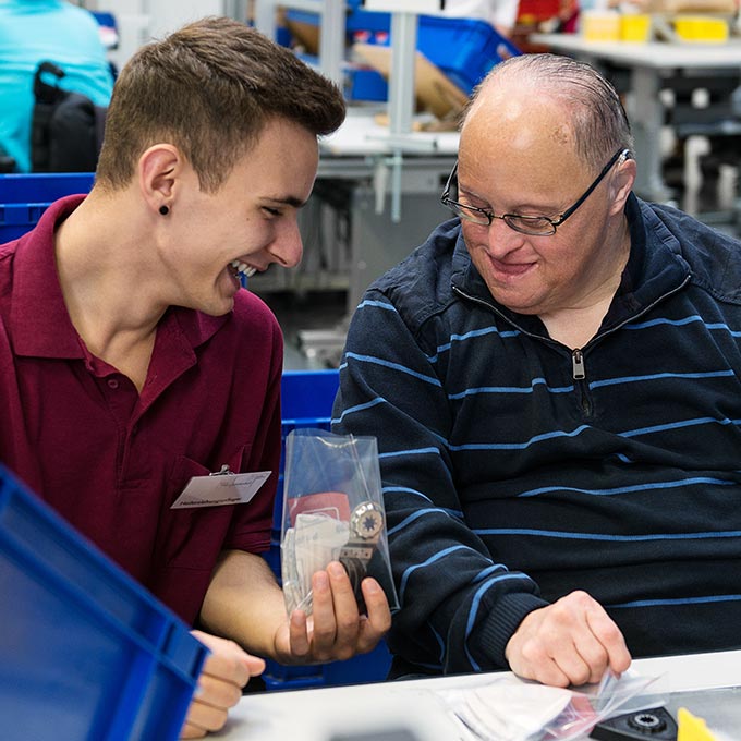 Ein junger Mann in einem kastanienbraunen Poloshirt und ein älterer Mann in einem gestreiften Pullover lächeln, während sie auf eine kleine Plastiktüte mit verschiedenen Kleinteilen blicken. Sie sitzen zwischen blauen Lagerbehältern in einer Werkstatt.