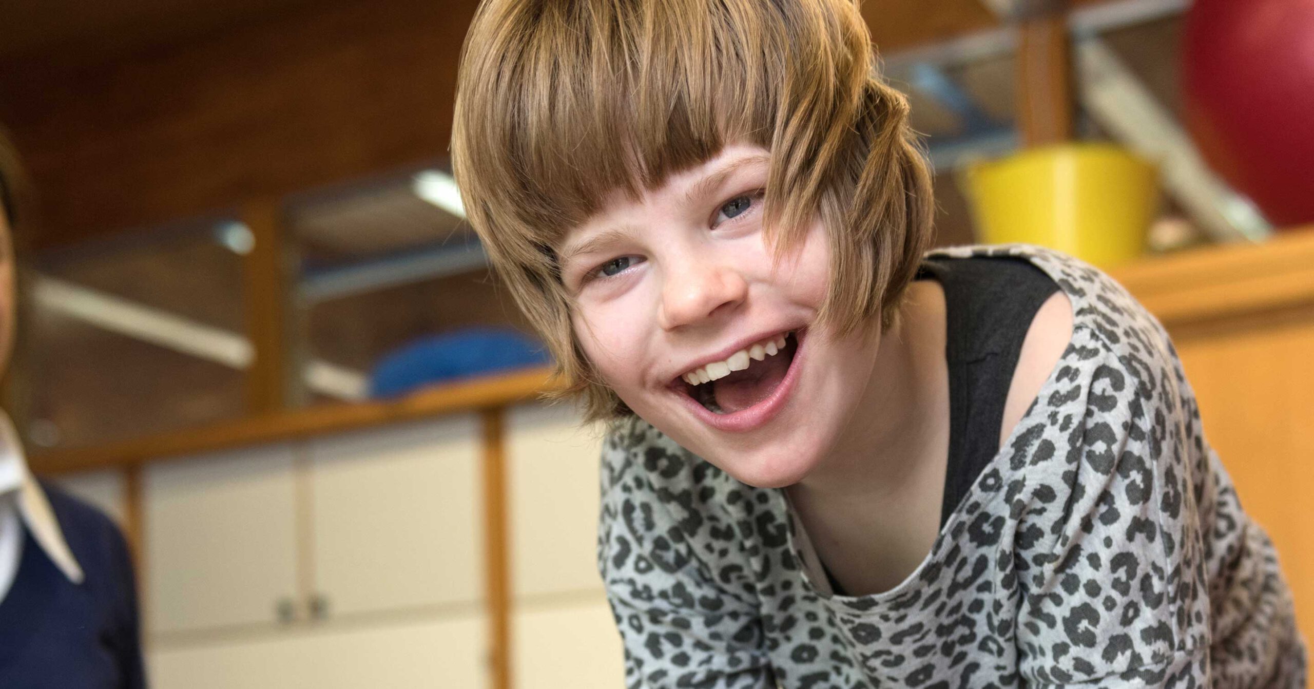 Ein kleines Kind mit kurzen braunen Haaren lächelt fröhlich in die Kamera und trägt ein Oberteil mit Leopardenmuster. Der Hintergrund mit Holzschränken und einem Teil eines roten Balls lässt die Wärme der Familie erahnen und erinnert an spielerische Tage nach der Schule.