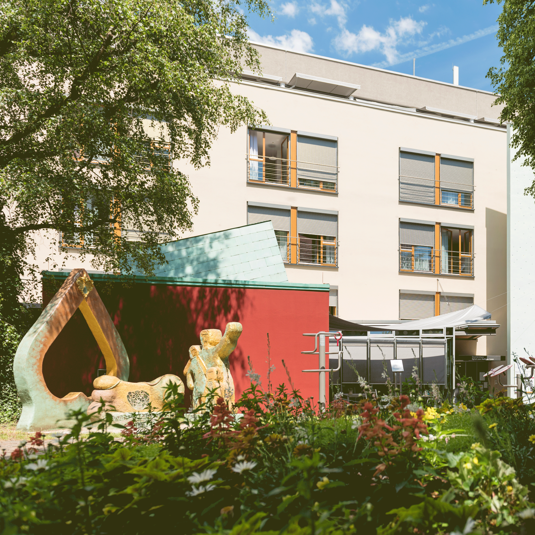 Hinter einem lebendigen Garten im historischen Heiligen Geist Spital steht ein modernes Gebäude mit großen Fenstern. Inmitten verschiedener Pflanzen und Blumen ruht eine farbenfrohe abstrakte Skulptur auf einer roten Plattform unter einem teilweise bewölkten blauen Himmel.