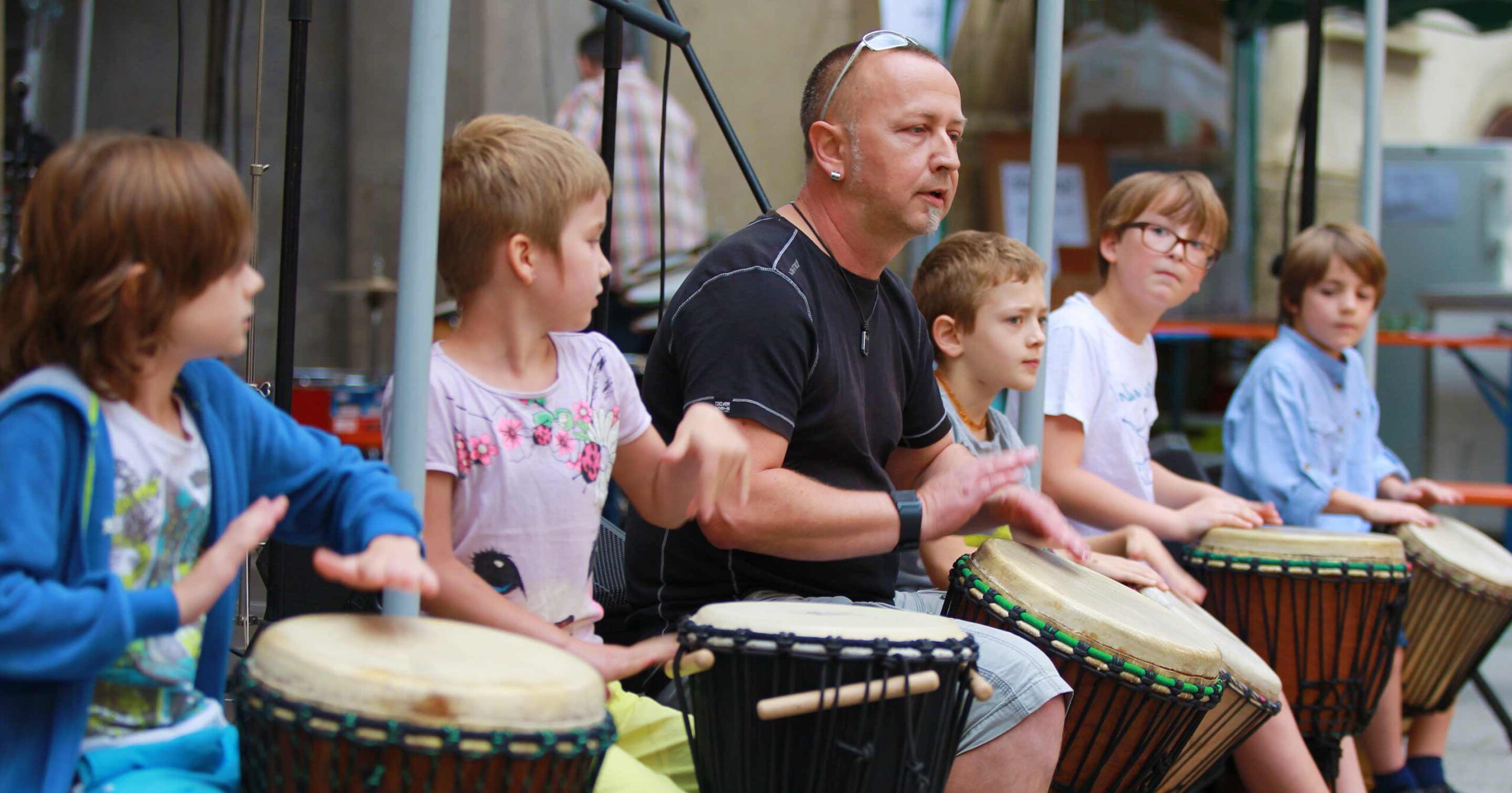 Eine Gruppe von Kindern und ein Erwachsener, einige davon mit Behinderung, sitzen in einer Reihe und spielen im Freien Handtrommeln. Sie wirken konzentriert und engagiert. Der Erwachsene steht in der Mitte und führt die Kinder zwischen verschiedenen Schlaginstrumenten hindurch.