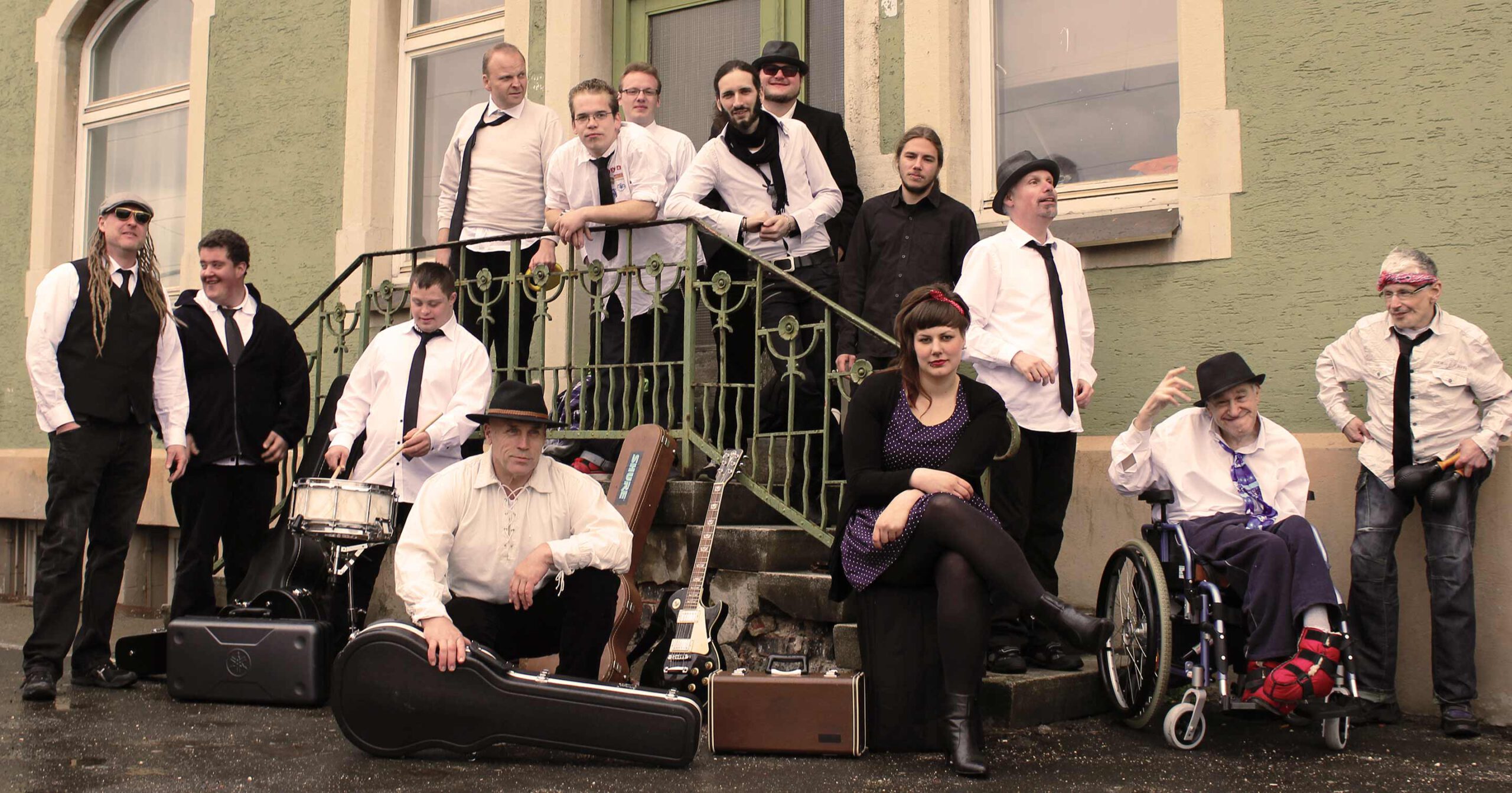 Eine Gruppe von Menschen, die überwiegend weiße Hemden und schwarze Krawatten tragen, posiert vor einem Gebäude. Eine Person, die Menschen mit Behinderung repräsentiert, sitzt in einem Rollstuhl, während andere Musikinstrumente wie Gitarren und eine Trompete halten. Die Szene hat ein Vintage-Gefühl mit gedämpften Farben.
