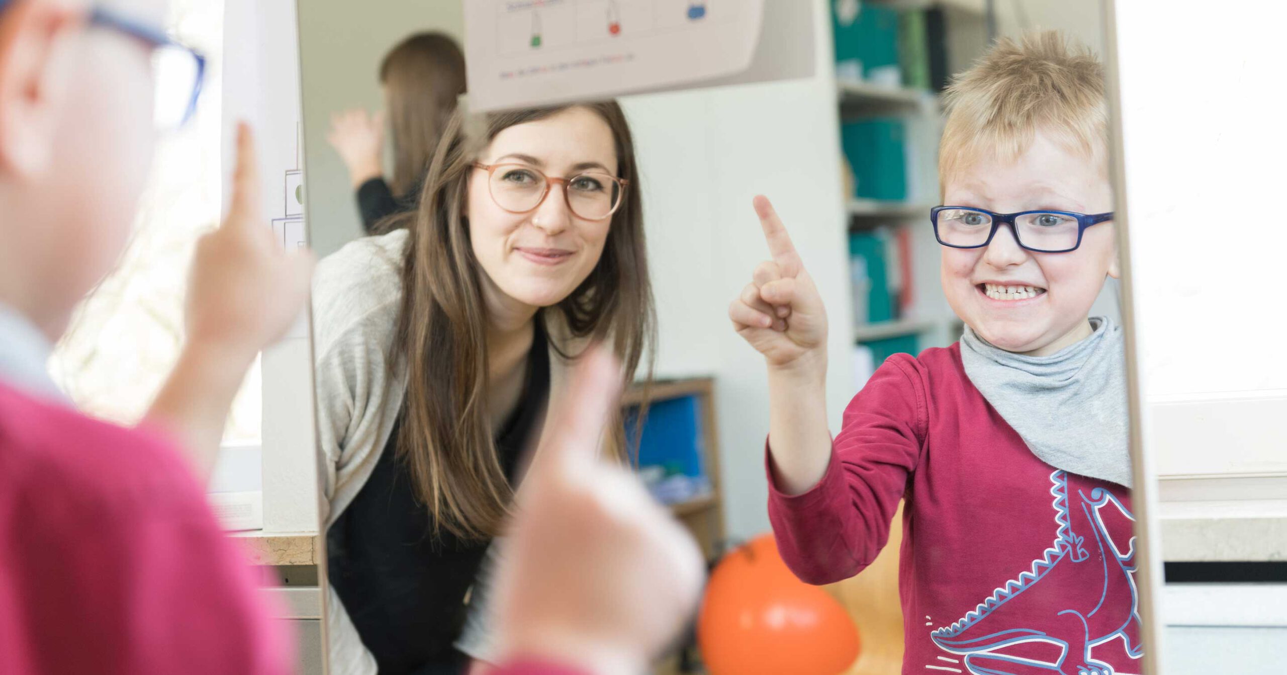 Ein Kind in rotem Hemd und Brille zeigt aufgeregt lächelnd auf einen Spiegel. Eine Frau mit Brille steht neben ihm und betrachtet lächelnd das Spiegelbild. Die Szene scheint ein heller und fröhlicher Raum zu sein.