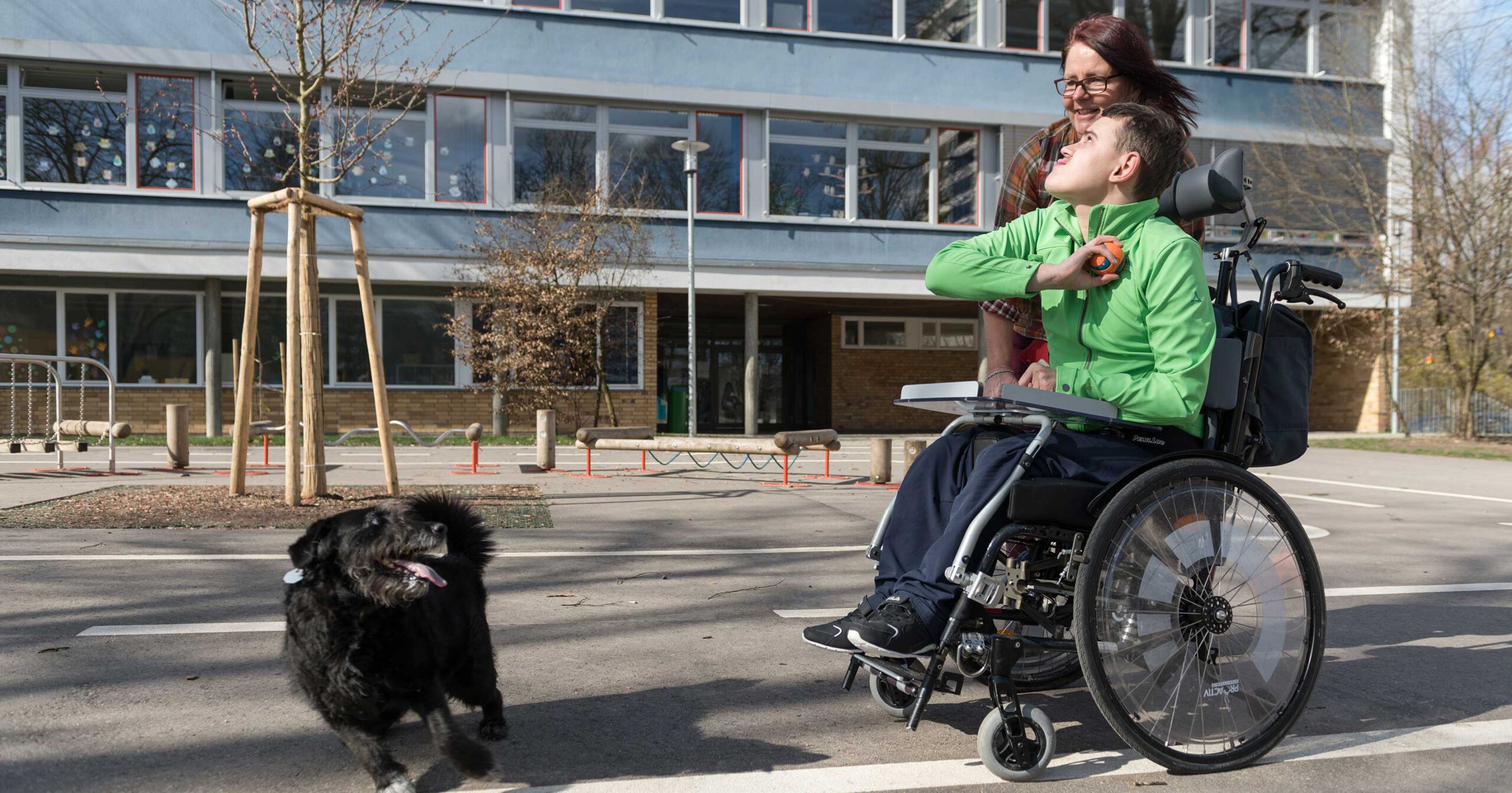 Ein junger Mensch mit schwerer Mehrfachbehinderung im Rollstuhl, der eine grüne Jacke trägt, lächelt und interagiert mit einem schwarzen Hund auf einem gepflasterten Weg. Ein Erwachsener steht in der Nähe und lächelt ebenfalls. Im Hintergrund sind ein Gebäude und kahle Bäume zu sehen.