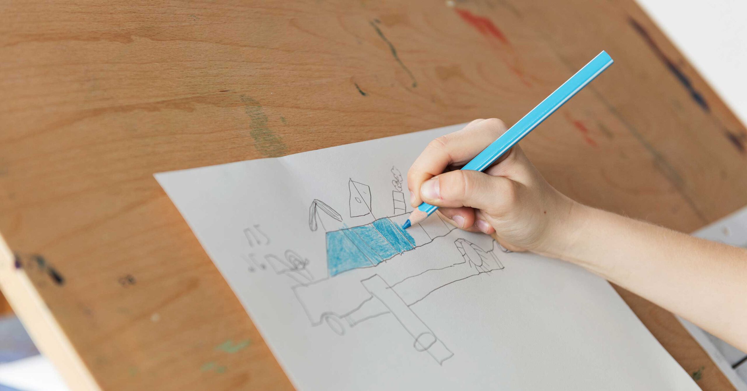 Ein Kind nimmt an Unterstützter Kommunikation teil, indem es mit einem blauen Buntstift auf weißem Papier zeichnet. Unter Führung der Kinderhand entstehen auf dem hölzernen Zeichenbrett abstrakte Formen und Linien.
