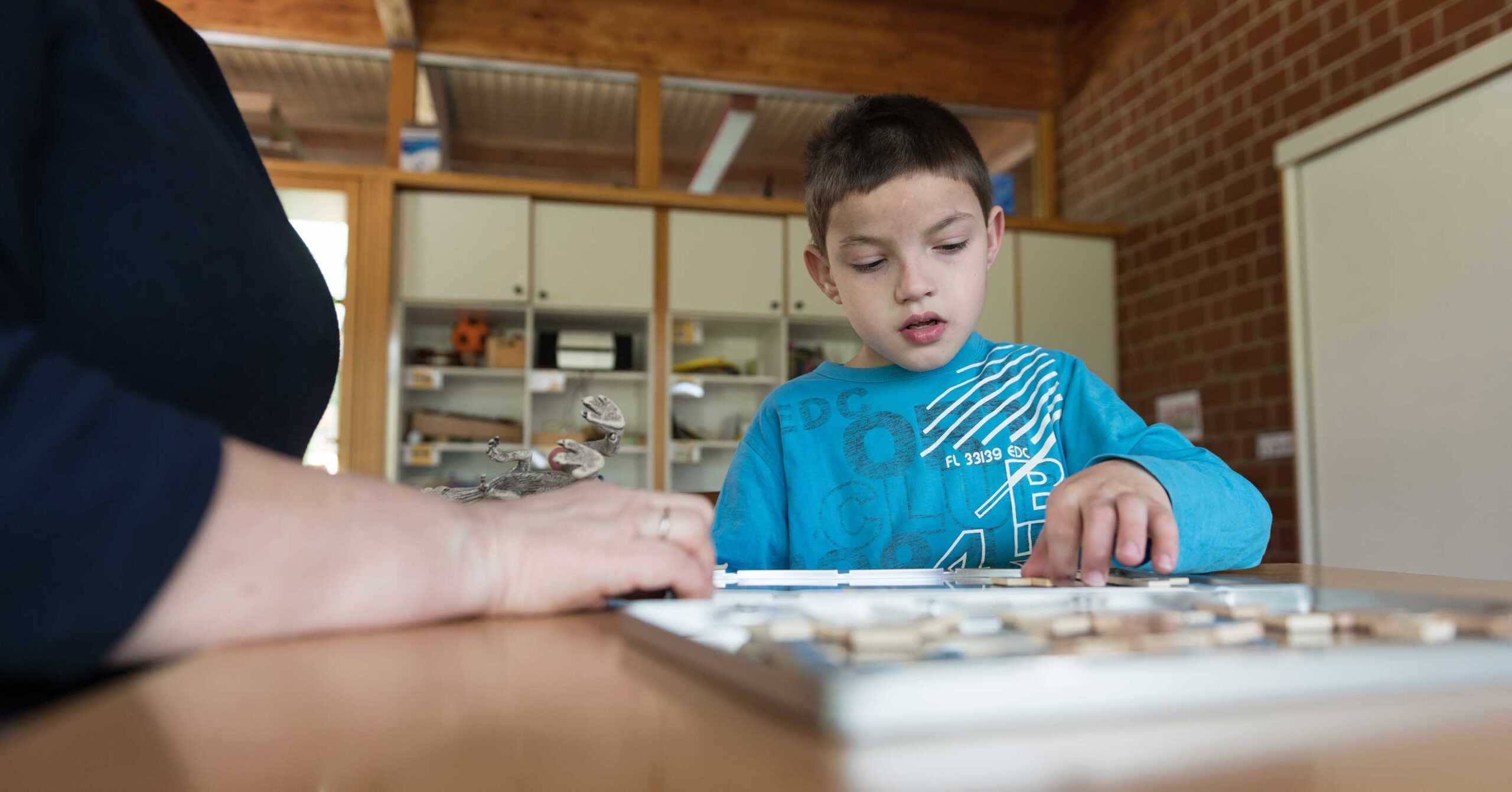 Ein kleiner Junge in einem blauen Hemd sitzt an einem Holztisch und konzentriert sich intensiv auf ein Brettspiel. Der Arm eines Erwachsenen ist seitlich zu sehen, verschwommen im Vordergrund, was an die strukturierte Flexibilität des Marchtaler Plans erinnert. Regale mit verschiedenen Gegenständen füllen den Hintergrund.