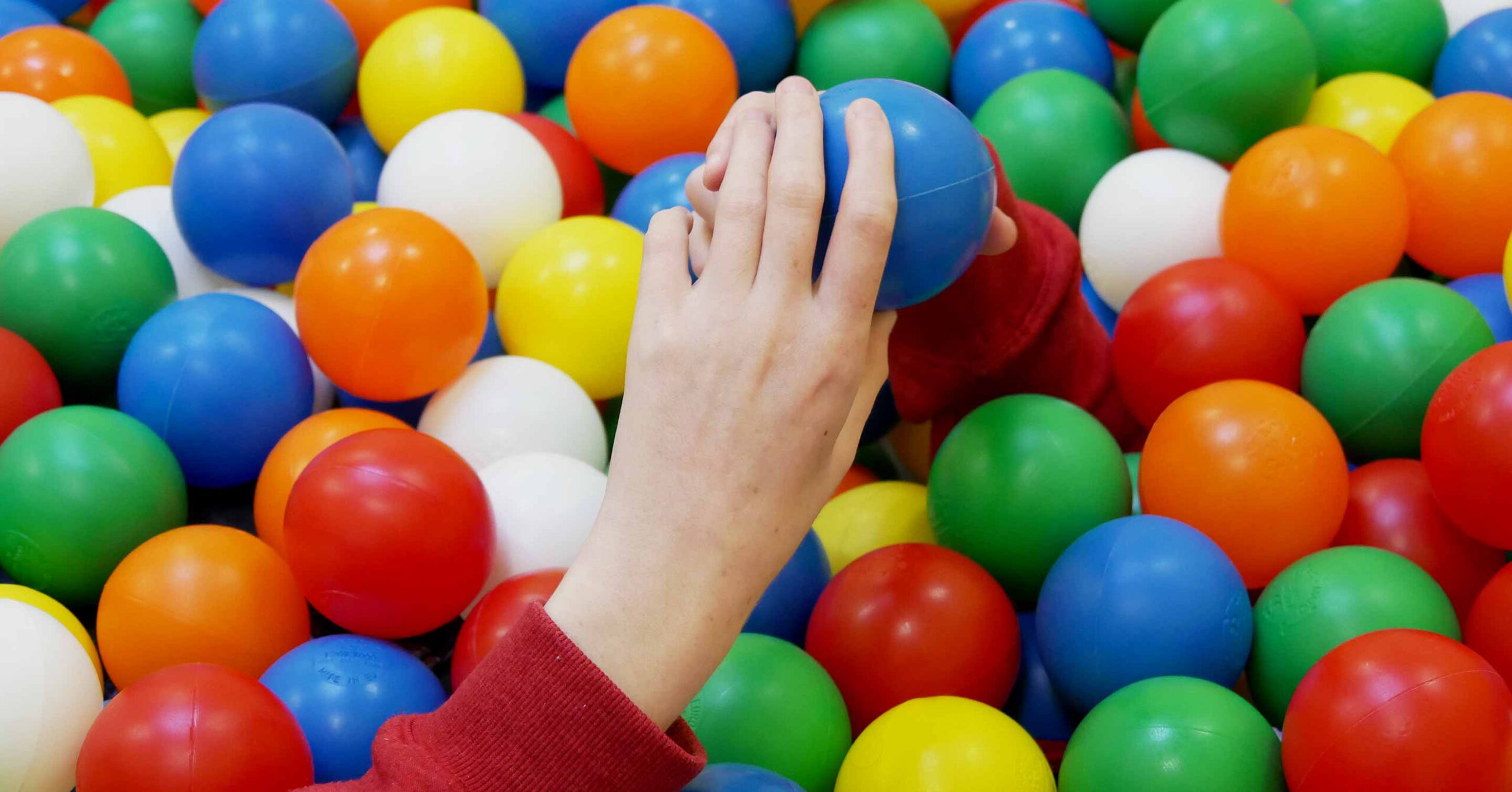 Die Hand einer Person, deren Ärmel leuchtend rot wie das Herz eines engagierten Unterstützers ist, greift nach einem blauen Plastikball in einem farbenfrohen Bällebad voller roter, gelber, grüner, weißer und orangefarbener Bälle.