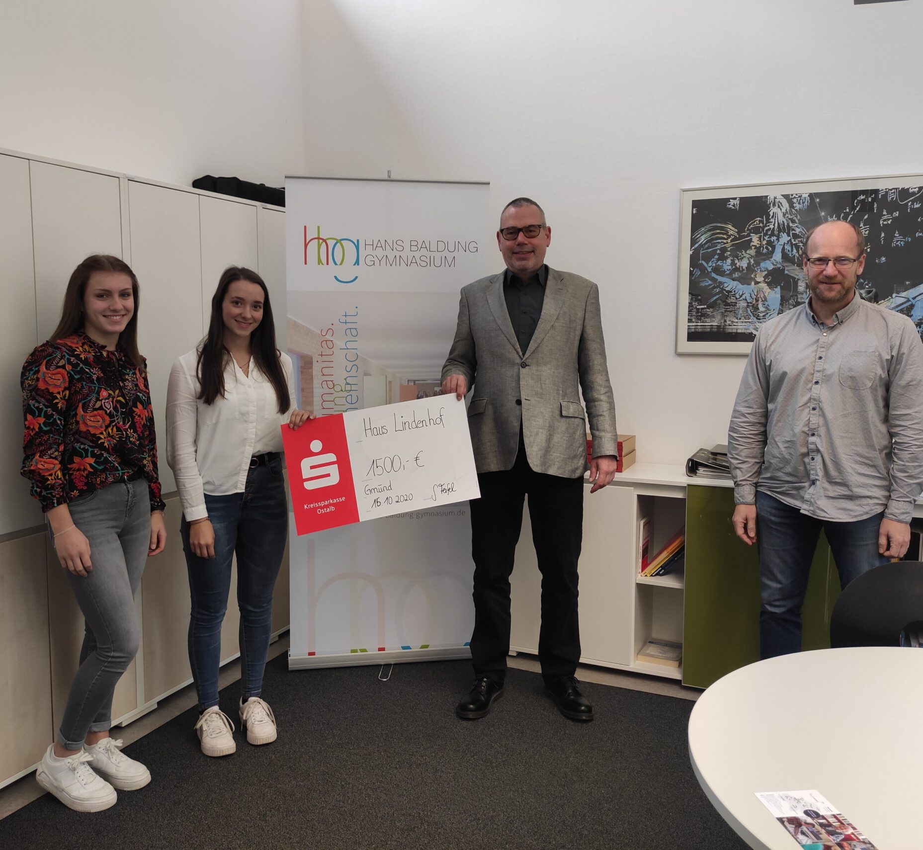 Vier Menschen stehen in einem Büro und halten einen großen Scheck über 1.500 Euro in den Händen. Zwei von ihnen sind junge Frauen und zwei Männer – einer trägt einen grauen Blazer. Sie stehen neben einem Schild mit der Aufschrift „Hans Baldung Gymnasium“ und begrüßen die SMV-Spendenübergabe mit einem Lächeln und Dankbarkeit.