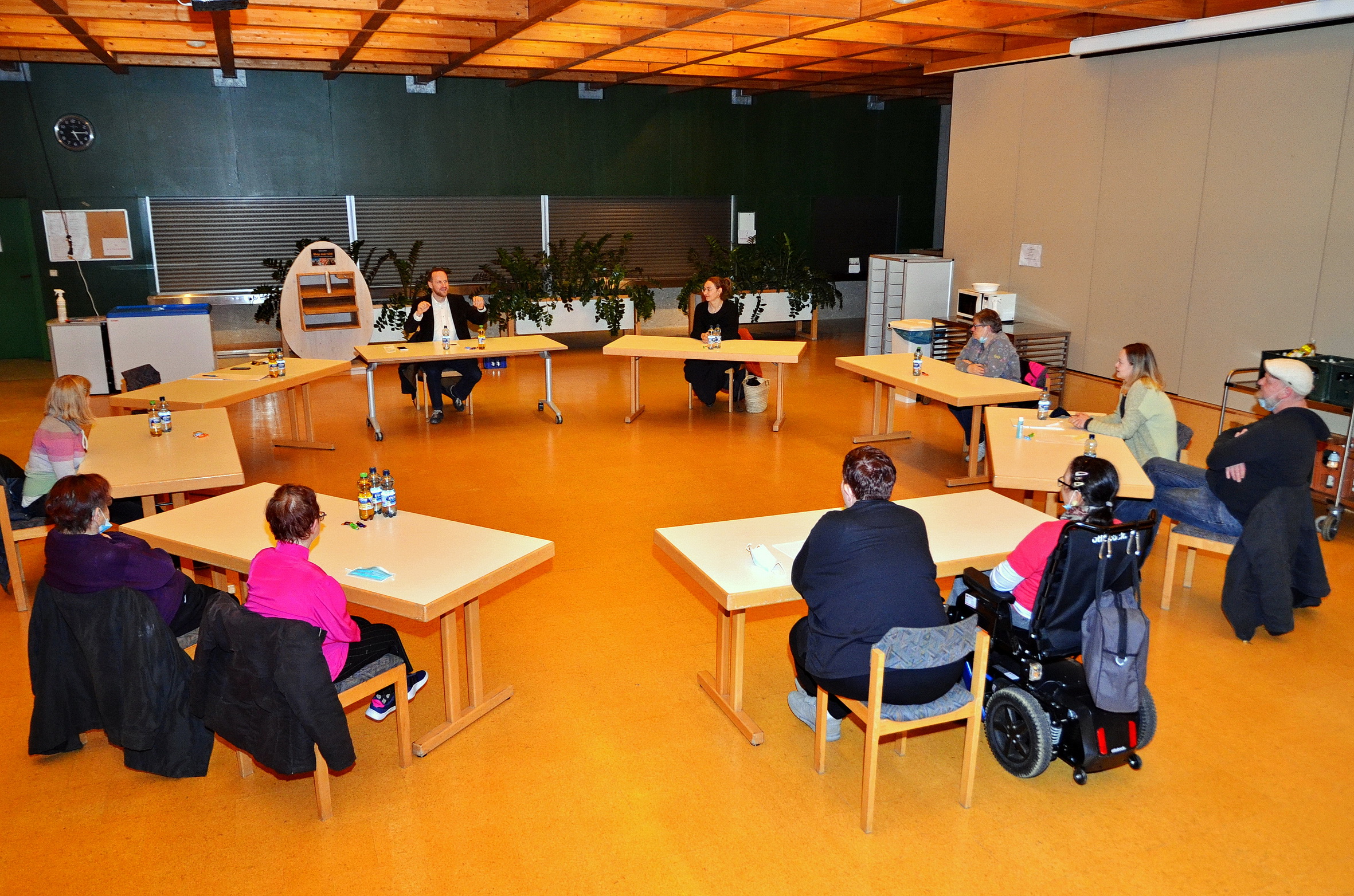 Eine Gruppe von zehn Personen sitzt um einen U-förmigen Tisch in einem Besprechungsraum mit gelbem Boden und Holzdecke. Der Bürgermeister leitet das Gespräch, während einige Personen Notizen machen, während andere aufmerksam zuhören. Rechts ist ein Rollstuhl zu sehen, der den Bedürfnissen aller gerecht wird.