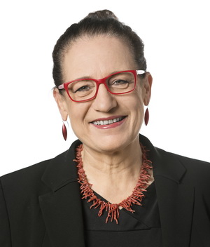 Eine Person lächelt herzlich und präsentiert ihre rote Brille, Halskette und Ohrringe. In einem eleganten schwarzen Oberteil und einer Jacke vor einem schlichten weißen Hintergrund strahlt sie das Selbstbewusstsein eines Mitglieds eines Kompetenzzentrums aus.