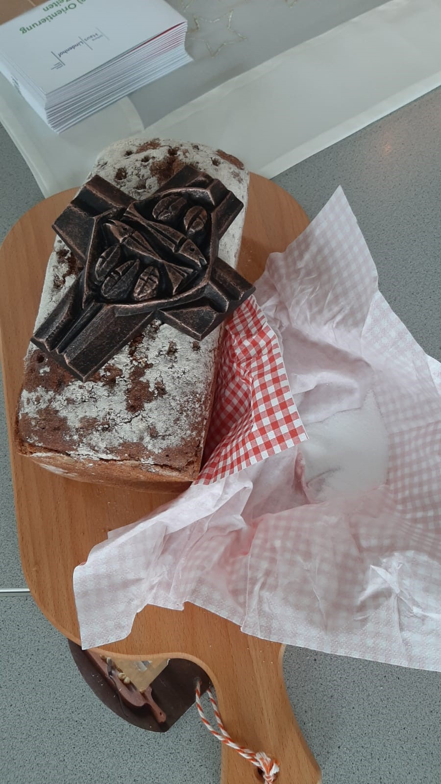 Ein Laib rustikales Brot mit einem dekorativen Schokoladenkreuz liegt auf einem hölzernen Schneidebrett. Das Brot ist teilweise aus dem rot-weiß karierten Papier ausgepackt und mit Puderzucker bestäubt, sodass es wahrhaft gesegnet aussieht. Im Hintergrund ist ein Stapel Papiere zu sehen.