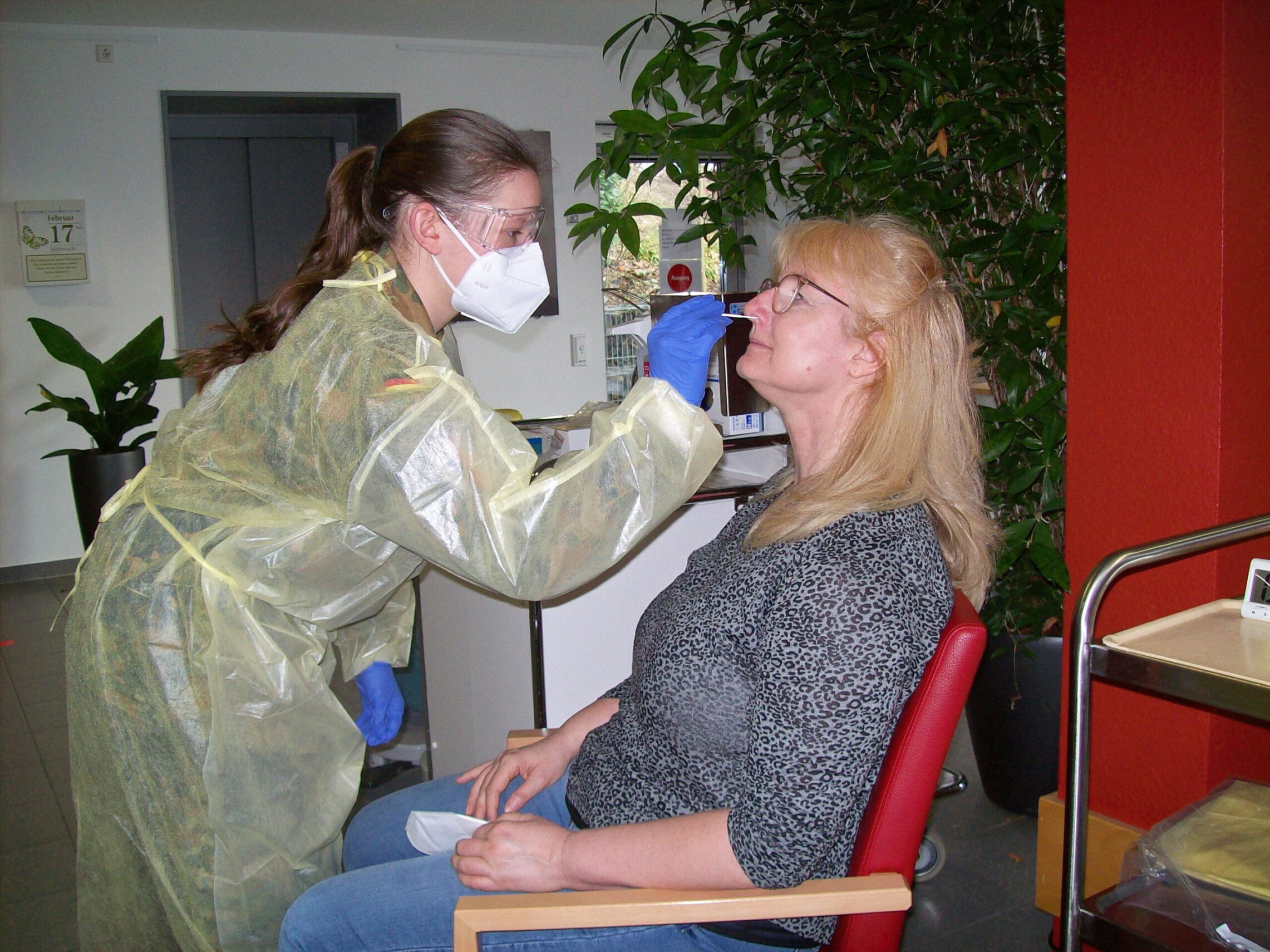 Ein Mitarbeiter des Gesundheitswesens führt in einer Klinikumgebung einen Nasenabstrichtest an einer sitzenden Frau durch. Die Frau, die ein gemustertes Oberteil trägt, sitzt ruhig da, während sich der Vorgang vor dem Hintergrund roter Wände und einer nahe gelegenen Pflanze abspielt.