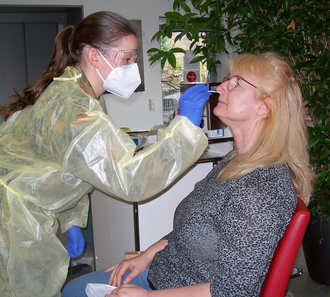 Ein Mitarbeiter des Gesundheitswesens in Schutzkleidung führt bei einer sitzenden Frau einen COVID-19-Test durch, indem er einen Nasenabstrich macht. Beide befinden sich im Innenbereich, wobei der Testende eine Gesichtsmaske, einen Gesichtsschutz und Handschuhe trägt. Tests werden weiterhin als wesentliche Dienste durchgeführt. Im Hintergrund ist eine grüne Pflanze zu sehen.