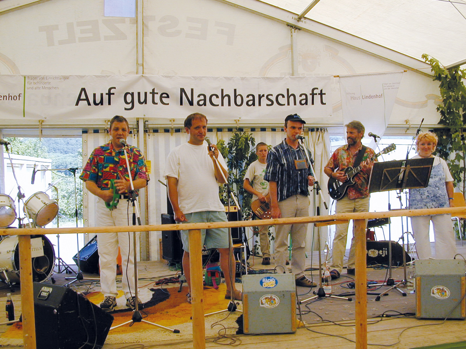 Eine Band tritt auf der Bühne unter einem Zelt mit dem Schild „Auf gute Nachbarschaft“ auf. Die sechs Mitglieder sind ein Schlagzeuger, ein Keyboarder und Gitarristen. Sie sind leger gekleidet und die Bühne ist mit dekorativem Grün umgeben.