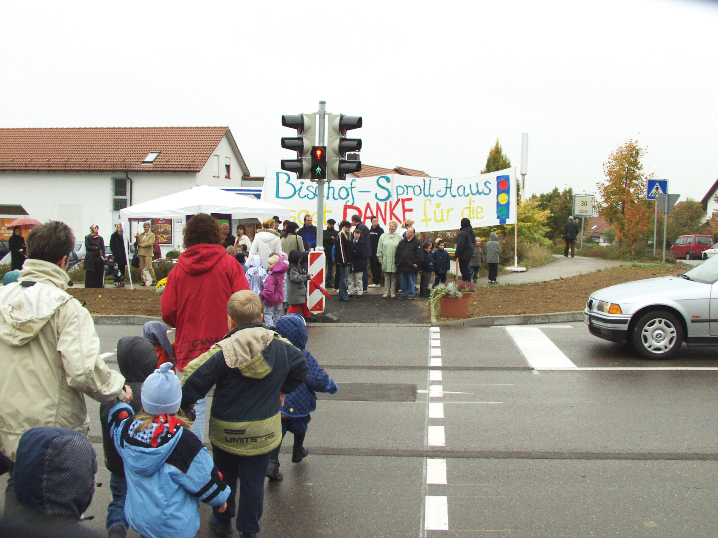 Eine Gruppe von Menschen, darunter auch Kinder, überquert eine Straße in Richtung einer Versammlung unter einem Zelt. Auf einem Banner steht auf Deutsch „Danke“. Ein silbernes Auto steht am Zebrastreifen. Es ist ein bewölkter Tag mit Häusern und Bäumen im Hintergrund.