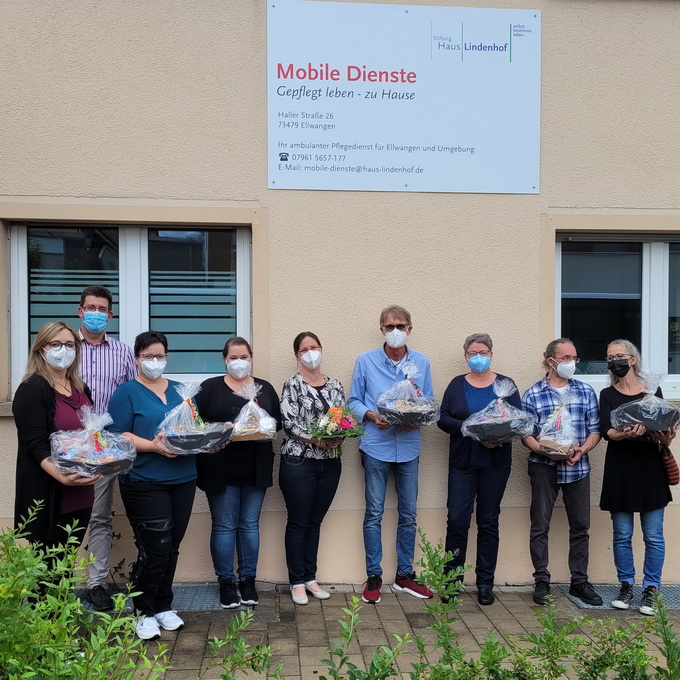 Eine Gruppe von neun Personen steht mit Geschenkkörben und einem Blumenstrauß vor einem Gebäude. Sie tragen Gesichtsmasken und Freizeitkleidung. Über ihnen steht ein Schild mit der Aufschrift „Mobile Task Force“, das das Ende ihrer Pflegemission markiert. Im Vordergrund sind Büsche zu sehen.