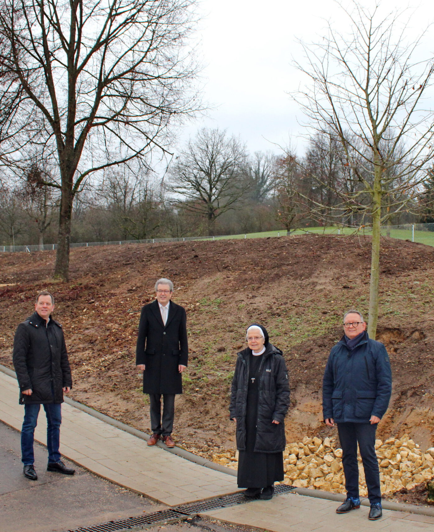 Vier Menschen stehen auf einem Gehsteig neben einem grasbewachsenen, kahlen Hügel. Im Hintergrund erstrecken sich blattlose und spärliche Linden unter einem bedeckten Himmel. Zwei Männer auf der linken Seite tragen dunkle Mäntel; in der Mitte steht eine Frau vom Klosterhospiz in einer Ordenstracht, während ein Mann auf der rechten Seite eine blaue Jacke trägt.