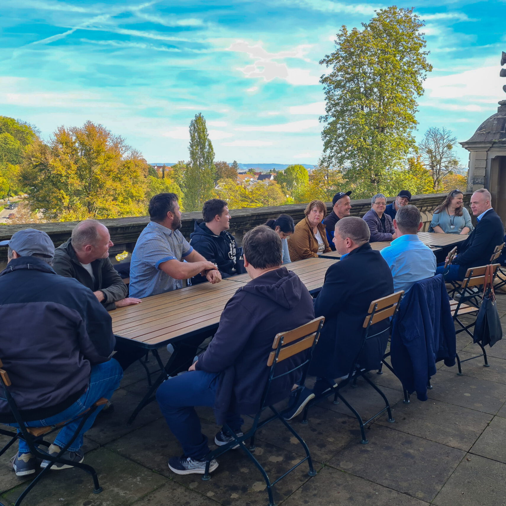 Eine Gruppe von Menschen sitzt draußen im Schlosshof um einen langen Holztisch herum und unterhält sich. Sie befinden sich auf einer Steinterrasse mit Bäumen und einer malerischen Landschaft im Hintergrund unter einem blauen Himmel mit dünnen Wolken.