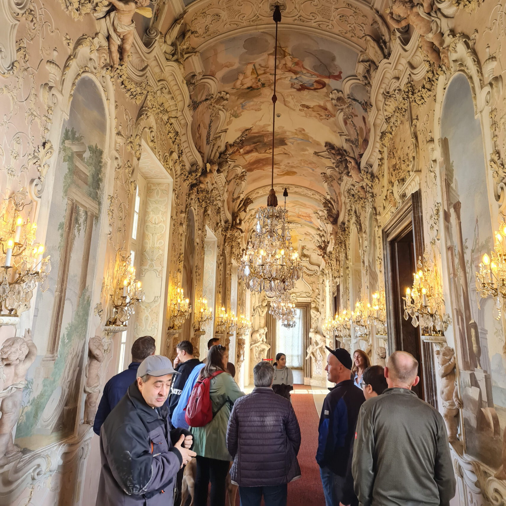 Eine Gruppe von Menschen besichtigt den reich verzierten Schlosshofflur, der mit aufwendigen Wand- und Deckengemälden im Barockstil, Kronleuchtern und Statuen geschmückt ist. Von Wandlampen beleuchtet, strahlt er eine warme, elegante Atmosphäre aus, die an ein Meisterwerk von Vinzenz von Paul-Werkstatt erinnert.