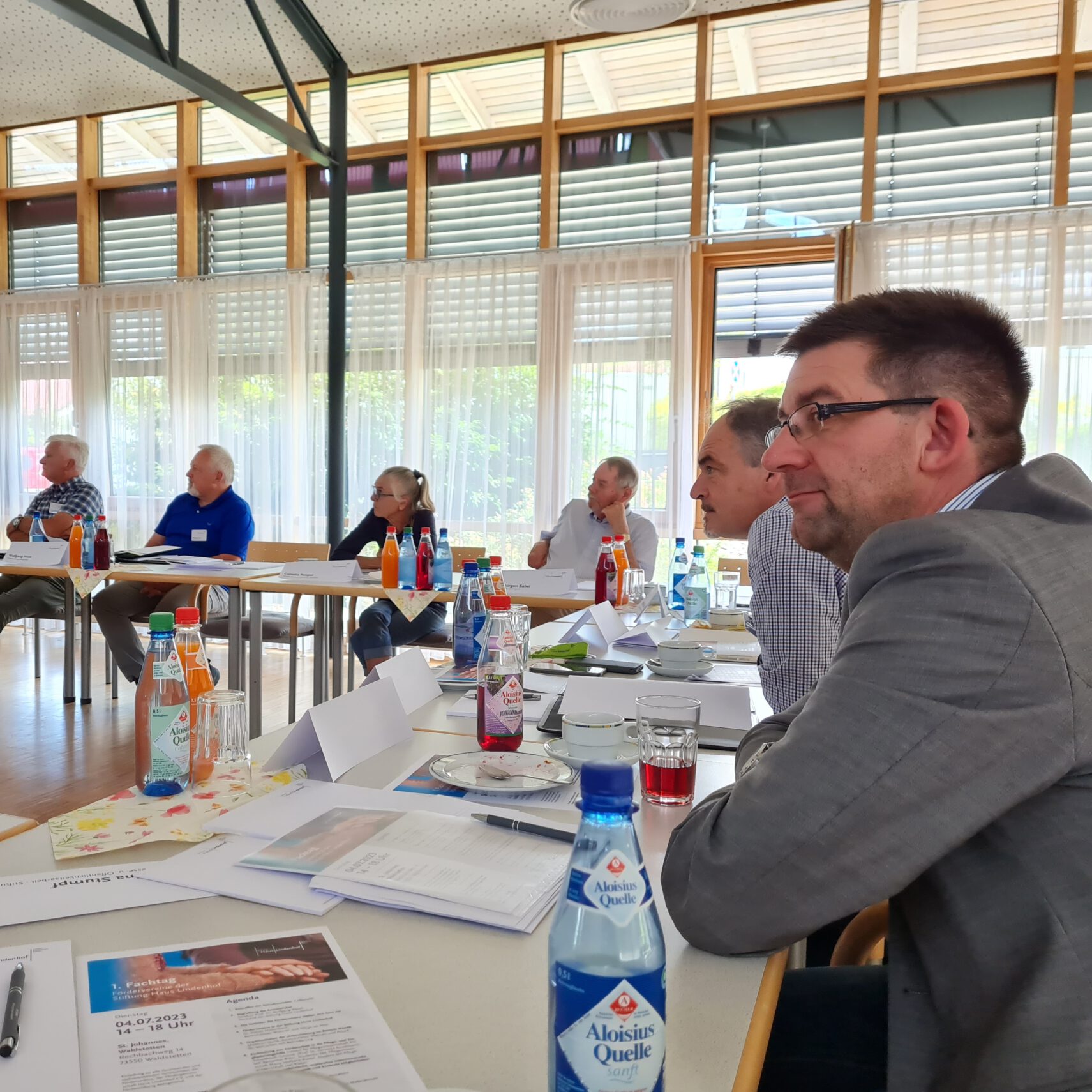 Eine Gruppe von Menschen sitzt an einem Konferenztisch in einem gut beleuchteten Raum und tauscht sich sinnvoll aus. Sie hören aufmerksam zu, vor ihnen liegen Dokumente, Namensschilder und Getränkeflaschen. Große Fenster mit transparenten Vorhängen lassen natürliches Licht in den Raum und schaffen so einen idealen Rahmen für dieses Treffen der Fördervereine.