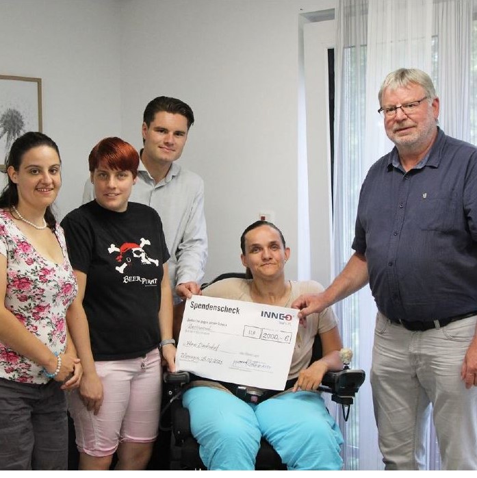 Eine Gruppe von fünf Personen hat sich in einem Innenbereich versammelt, eine Frau im Rollstuhl hält einen großen Spendenscheck über 2.000,00 Euro der Stiftung Haus Lindenhof in den Händen. Sie lächeln in die Kamera in Ellwangen. Im Hintergrund ist ein großes Fenster mit transparenten Vorhängen zu sehen.