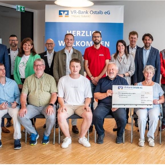 Eine Gruppe von Menschen posiert drinnen mit einem großen Festscheck der VR-Bank-Stiftung. Einige sitzen, andere stehen im Hintergrund. Auf einem Schild hinter ihnen steht „VR-Bank Ostalb eG“ und „Herzlich Willkommen“. Lächelnd in Freizeit- und Businesskleidung feiern sie die erfolgreiche Ausschüttung.