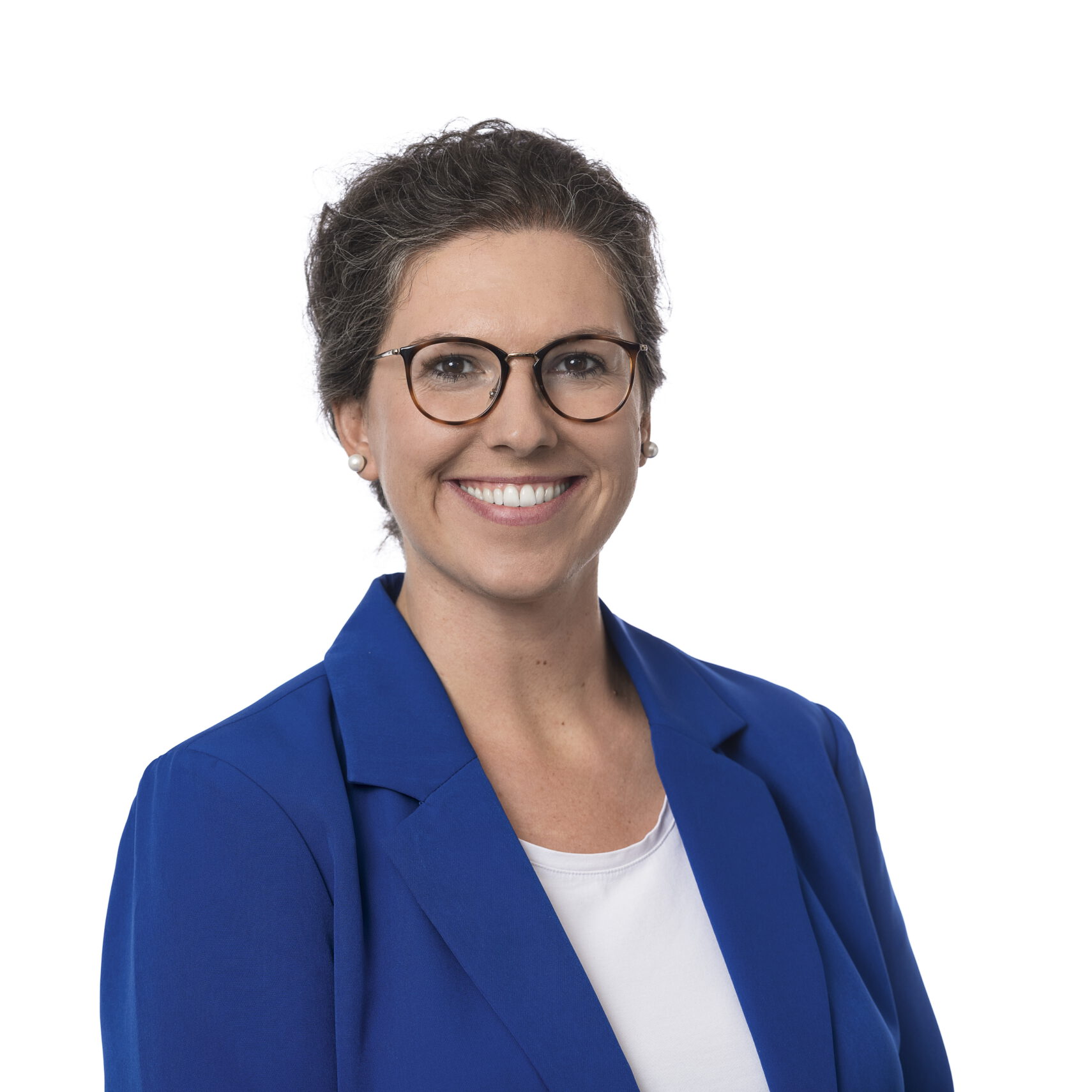 Eine Frau mit kurzen Haaren und Brille lächelt und trägt einen blauen Blazer über einem weißen Hemd. Sie posiert vor einem schlichten weißen Hintergrund.