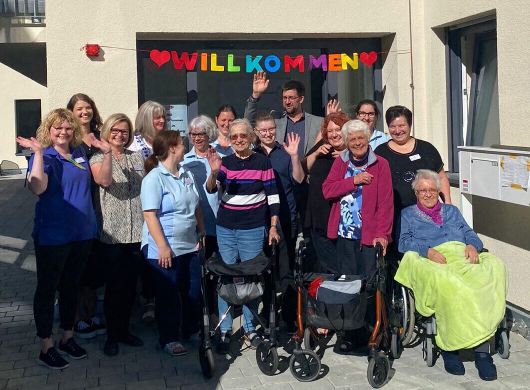 Eine Gruppe von Menschen, darunter ältere Menschen und Pflegekräfte, versammelt sich freudig am Eingang von St. Georg Steinheim unter einem „WILLKOMMEN“-Banner. Einige sitzen in Rollstühlen, andere winken oder lächeln und feiern den Einzug der ersten Bewohner an diesem sonnigen Tag.