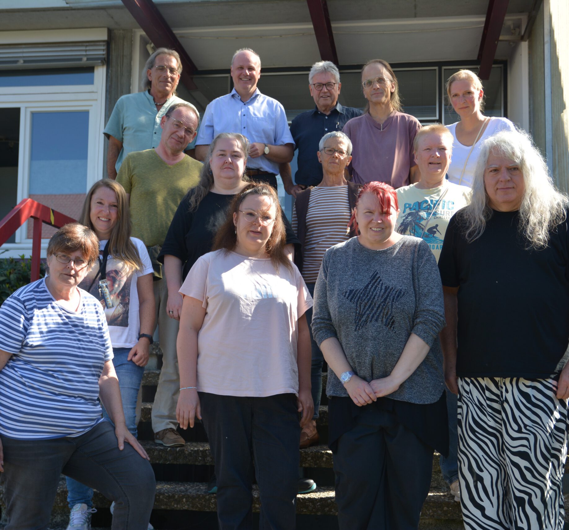 Eine Gruppe von vierzehn Personen steht auf einer Außentreppe vor einem Gebäude und posiert für ein Foto im Rahmen des Projekts PRODI-Werkstatt. Die bunt gemischte Gruppe ist leger gekleidet und lächelt. Sie besteht aus Männern und Frauen, einige tragen Brillen.