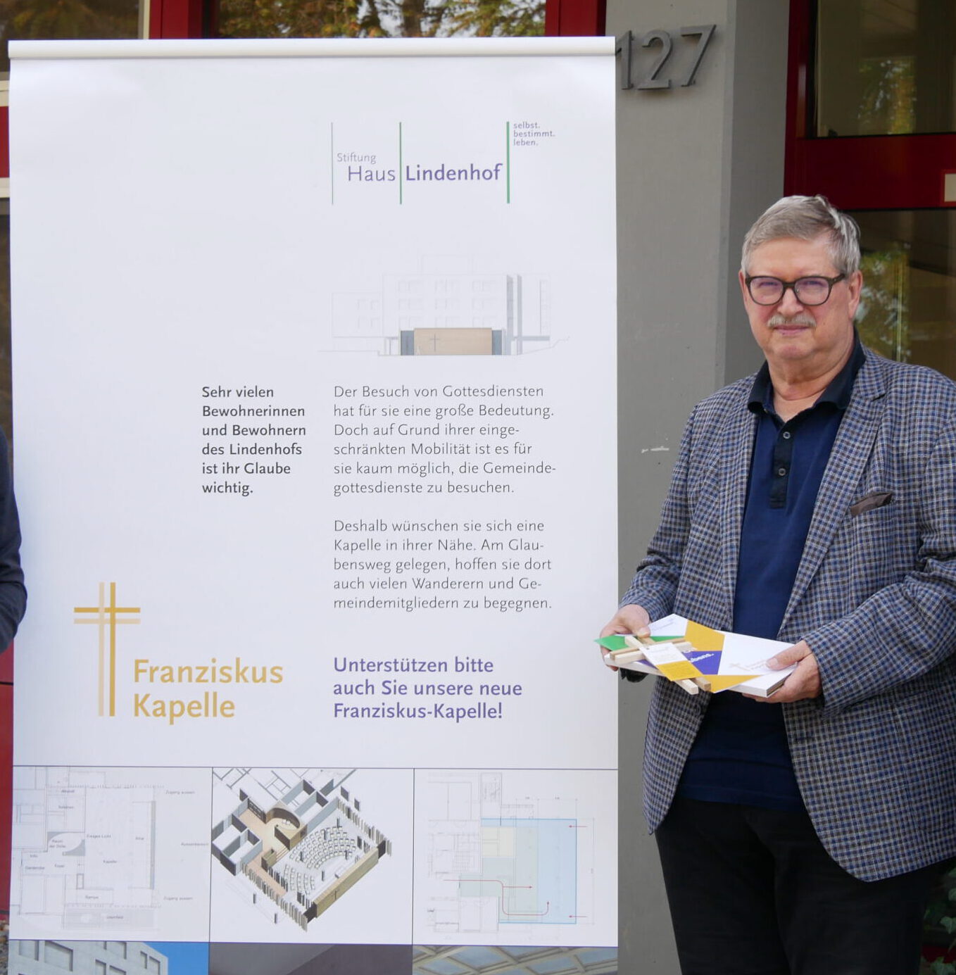 Eine Person steht neben einer Schautafel mit Bauplänen für die Franziskus-Kapelle, die vermutlich von Jürgen Kunze kuratiert wurde. Sie hält Broschüren in der Hand, trägt eine Brille und ist leger gekleidet. Die Tafel unterstreicht die Bedeutung der Kapelle für die örtliche Gemeinde.