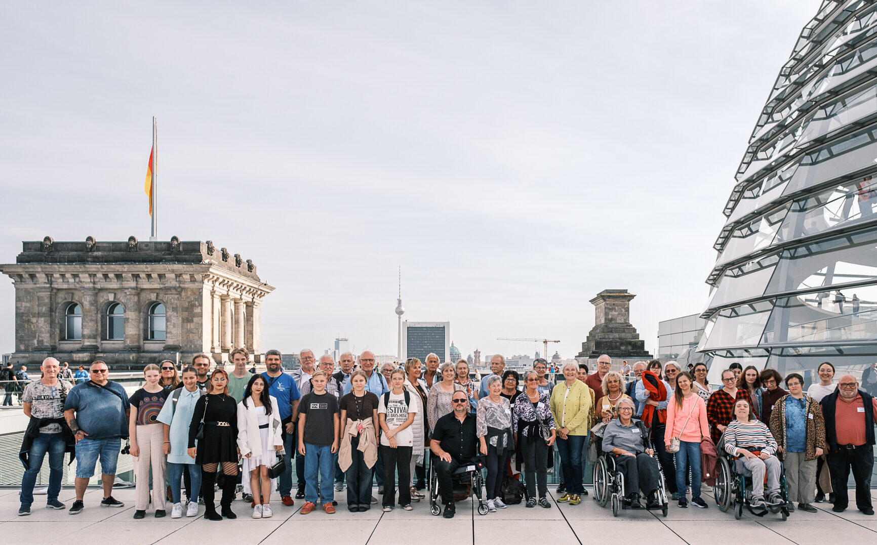 Eine große Gruppe von Menschen steht auf einem Dach in Berlin, im Hintergrund zwei historische Gebäude und eine Glaskuppel. Der klare Himmel hebt die deutsche Flagge hervor, die auf einem der Gebäude weht. Dies schafft die Kulisse für eine bereichernde Bildungsfahrt mit Schwerpunkt auf politischer Bildung.