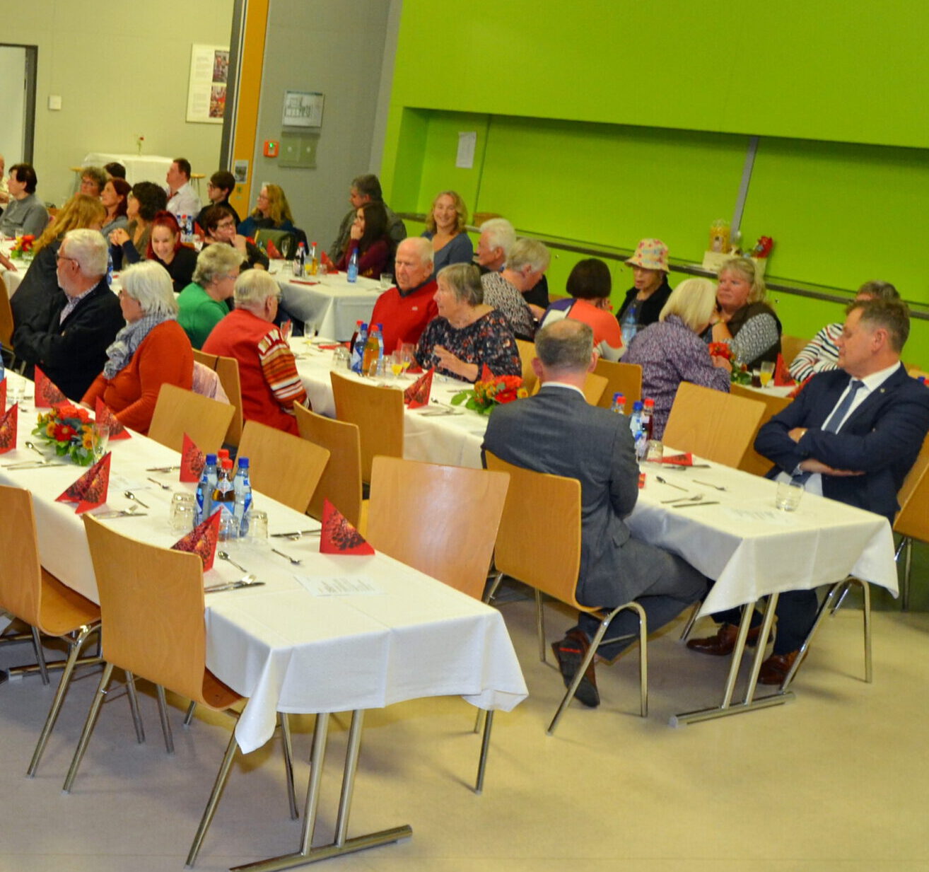 In einem hell erleuchteten Raum mit grünen Wänden sitzt eine Gruppe von Menschen an Tischen und feiert ein Ehrenamtsfest. Auf den mit weißen Tischdecken und roten Servietten geschmückten Tischen liegen kleine Blumenarrangements als Zeichen der Anerkennung und Dankbarkeit. Die Menschen unterhalten sich angeregt.