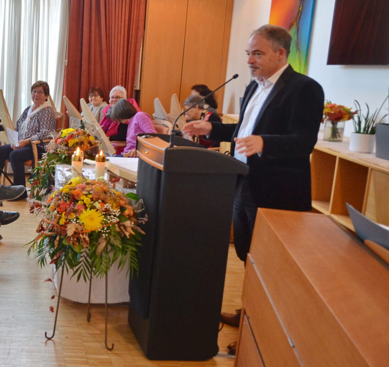 Ein Mann im Anzug spricht an einem Podium zu den Teilnehmern der St. Lukas-Kirche in Abtsgmünd. Der mit herbstlichen Blumenarrangements geschmückte Raum strahlt Wärme und Schönheit aus. Während sich einige Teilnehmer Notizen machen, verleiht das farbenfrohe Kunstwerk hinter dem Podium dieser Versammlung im Pflegeheim einen Hauch von Glanz.