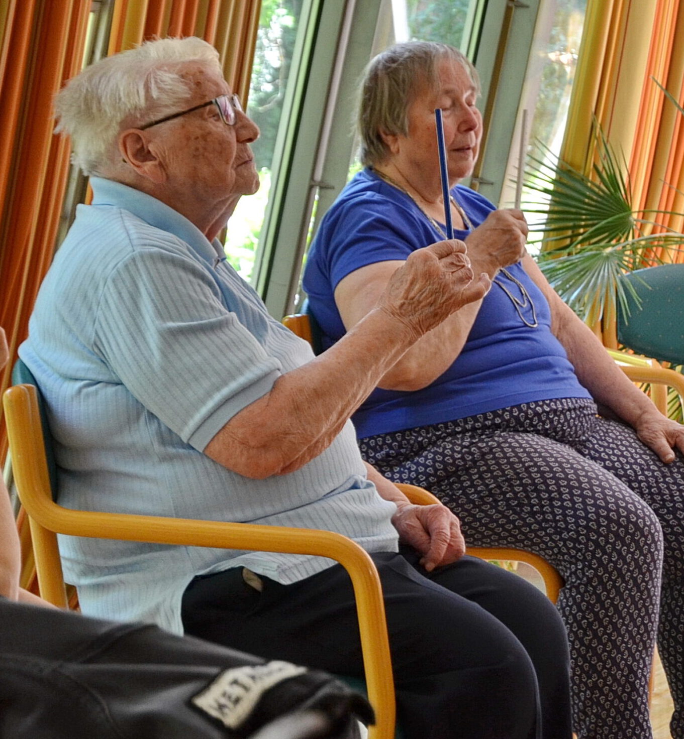 Ein älterer Mann und eine ältere Frau sitzen nebeneinander und üben eine Indoor-REHA-Sportaktivität aus. Jeder hält einen nach oben gerichteten Bleistift, konzentriert und konzentriert. Der Raum ist gut beleuchtet, mit bunten Vorhängen und Grünpflanzen im Hintergrund, was an eine ruhige Sitzung im Spital Hl. Geist erinnert.