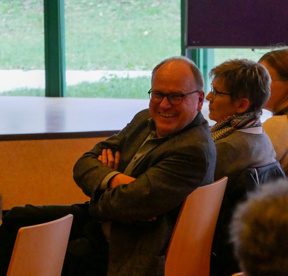 Michael Abele, mit Brille und grauem Blazer, lächelt warm in die Kamera, während er neben einer kurzhaarigen Frau mit Kopftuch sitzt. Hinter ihnen geben grüne Fenster einen Blick auf das üppige Gras draußen frei. Sie sind Teil der Stiftung Haus Lindenhof, die ihr Erbe über 40 Jahre würdigt.