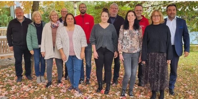 Eine Gruppe von zwölf Personen steht gemeinsam im Freien auf einer mit Herbstlaub geschmückten Rasenfläche und verkörpert den Geist von selbst.bestimmt.leben. Sie sind in legerer und halbformeller Kleidung gekleidet, umrahmt von Bäumen mit leuchtendem Herbstlaub, was die Werte widerspiegelt, die von der Abtsgmünder Förderstiftung unterstützt werden.