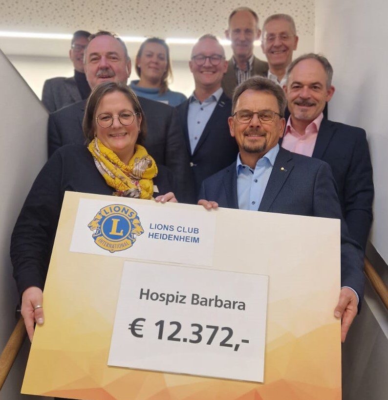 Strahlend steht eine Gruppe von neun Personen auf einer Treppe und überreicht einen großen Spendenscheck des LIONS Clubs Heidenheim. Die großzügige Spendenübergabe in Höhe von 12.372 Euro ist für das Hospiz Barbara bestimmt.