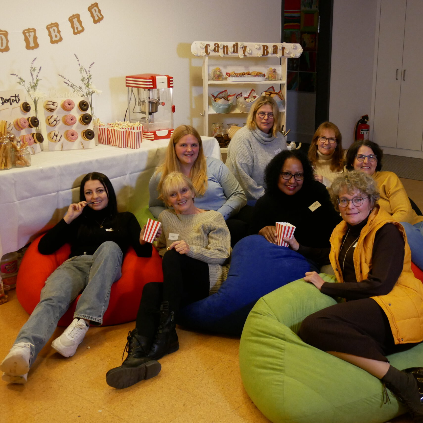 Eine Gruppe von neun Personen lächelt auf bunten Sitzsäcken sitzend in die Kamera und fängt den perfekten Social-Media-Moment ein. Hinter ihnen befindet sich eine Candybar mit Popcorn, Donuts und Süßigkeiten. Der Raum strahlt eine fröhliche, lockere Atmosphäre aus und macht das Ganze zu einem unvergesslichen Event.