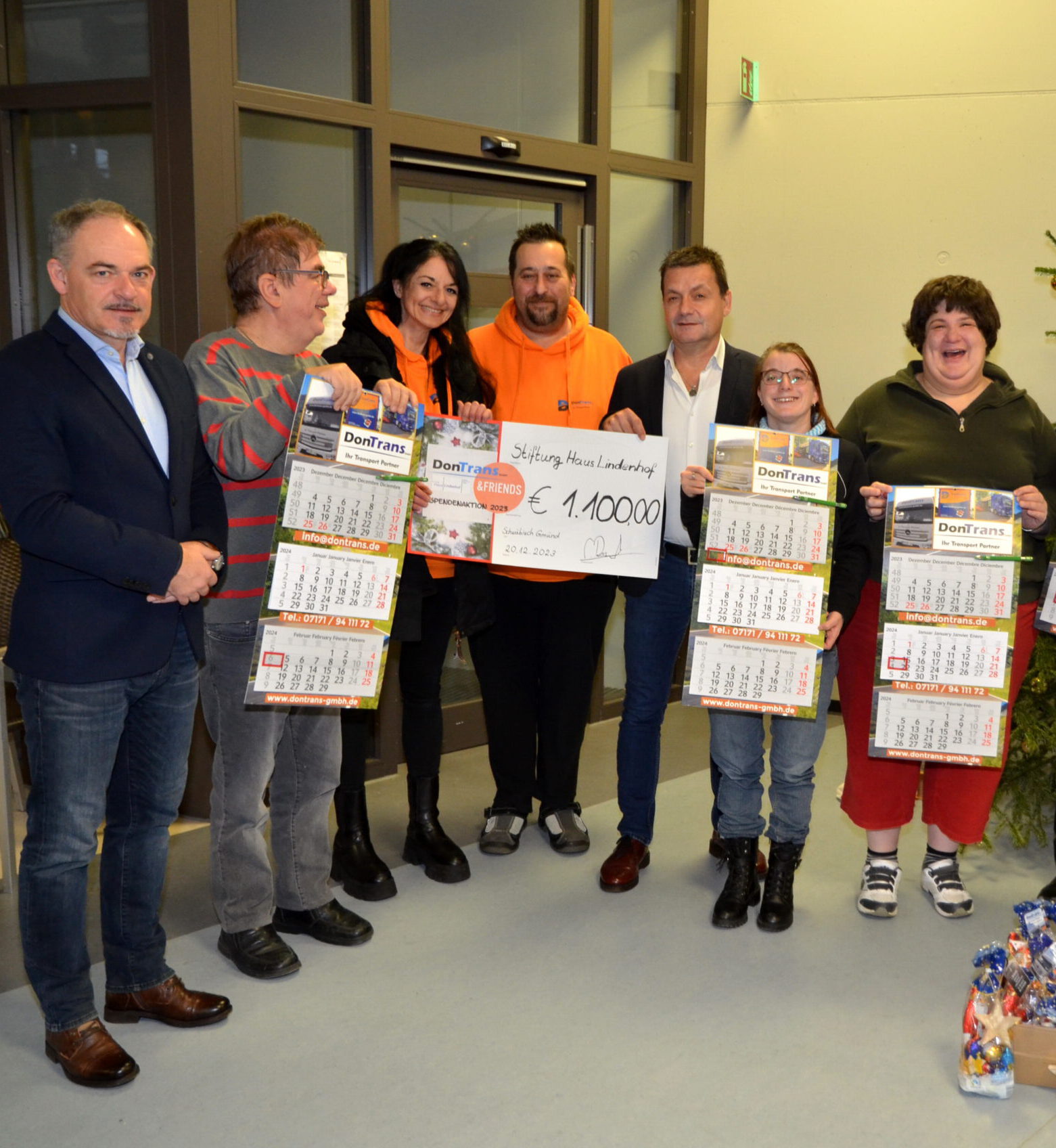 Sieben Personen, darunter Freunde der DonTrans GmbH, stehen lächelnd mit Kalendern und einem großen Scheck über 1.000 Euro im Haus. Die Gruppe, in Freizeit- und Businesskleidung gekleidet, scheint sich in einem informellen Rahmen zu befinden und ihre erfolgreiche Spendenaktion zu feiern.