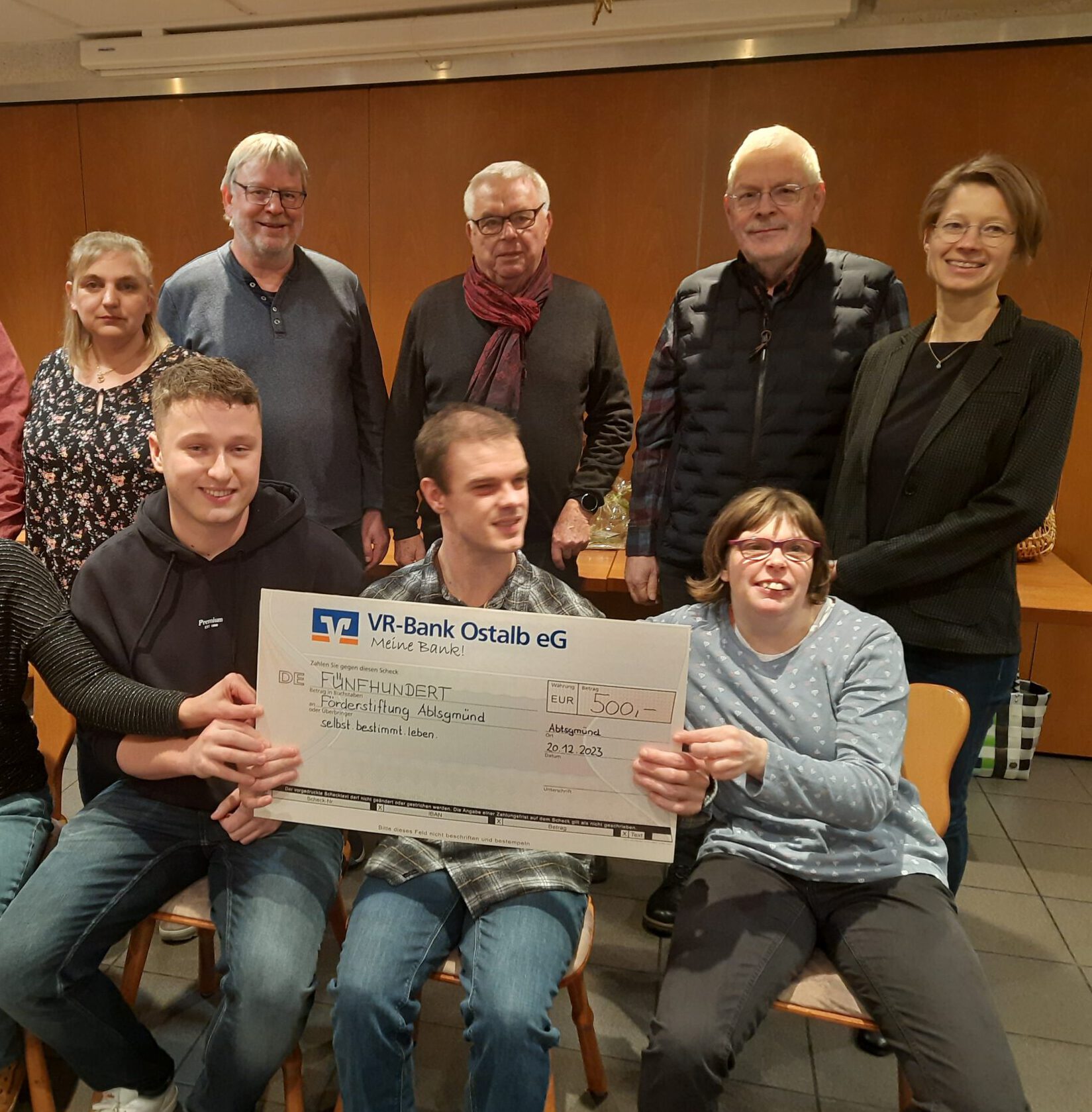 Eine Gruppe sitzender und stehender Menschen vom Ökumenischen KirchenChor in Abtsgmünd posiert glücklich mit einem großen Scheck über 500 Euro. Der Scheck ist eine großzügige Spende der VR-Bank Ostalb eG vom 24.02.2023. Alle lächeln in dem warm beleuchteten Raum.