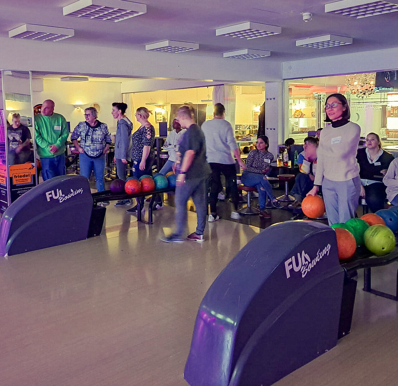 In einer gut beleuchteten Bowlinghalle versammeln sich Azubi-Bowling-Teilnehmer mit bunten Bällen um die Bahnen. Einige schauen gespannt zu, während andere sich zum Bowlen bereit machen. Währenddessen bieten ehrenamtliche Helfer der Altenhilfe freundliche Unterstützung von den Sitzbereichen aus, umgeben von glänzender Bowlingausrüstung.