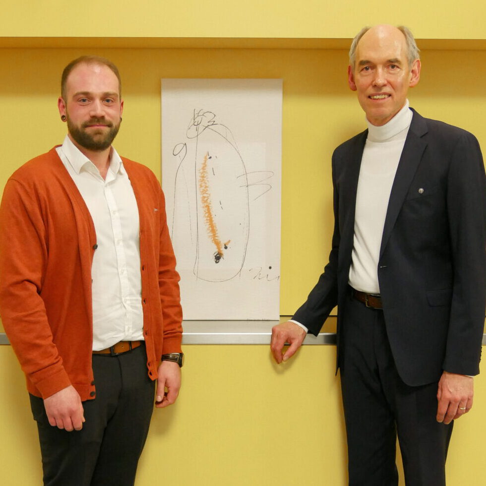 Zwei Männer stehen vor einer gelben Wand im Bischof Ketteler Haus, zwischen ihnen eine Zeichnung. Der Mann links trägt ein weißes Hemd und eine rote Strickjacke, während der Mann rechts einen marineblauen Anzug und einen weißen Rollkragen trägt. Das abstrakte Kunstwerk spiegelt den dynamischen Leitungswechsel wider, der darin stattfindet.