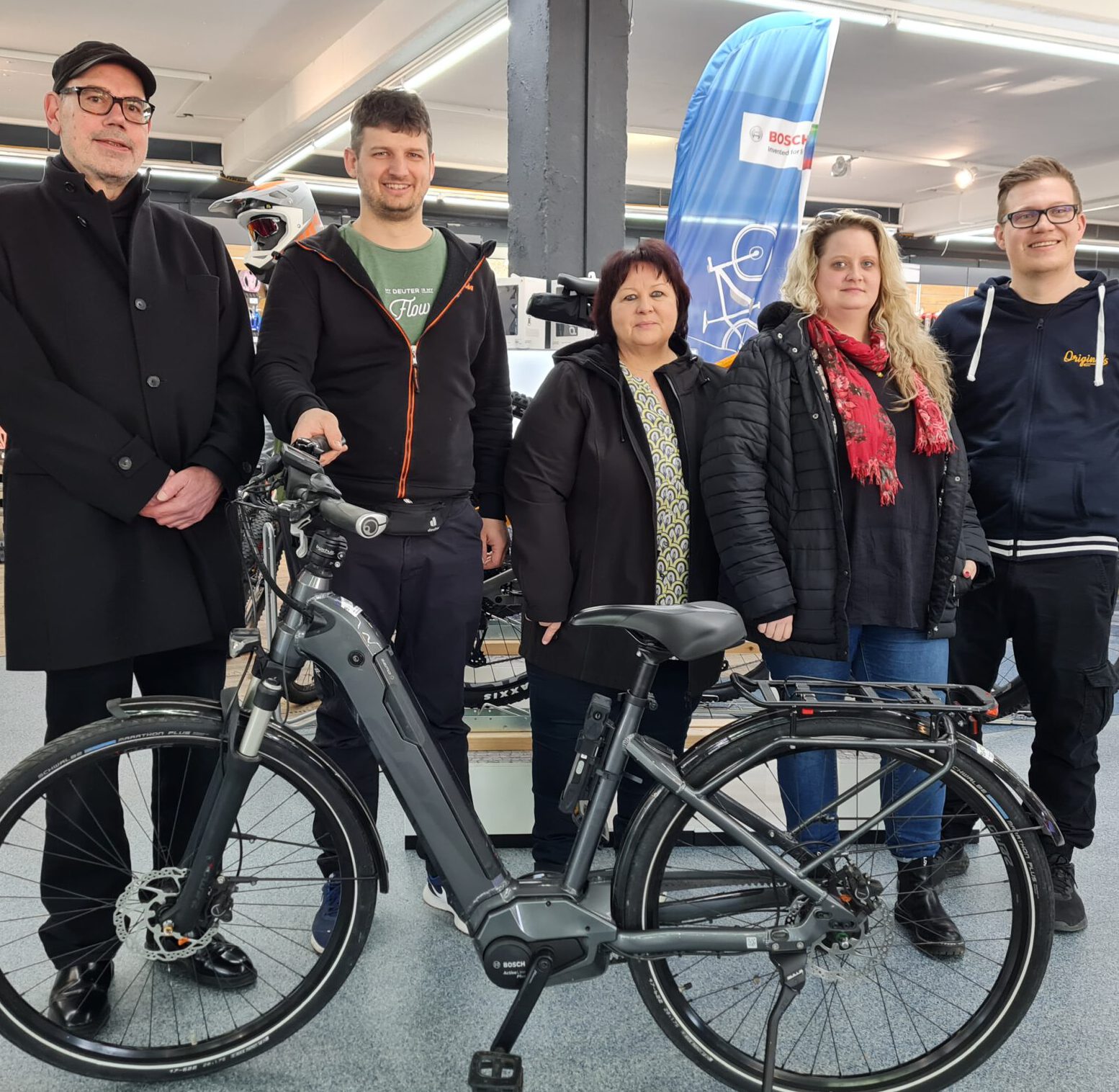 Im Ausstellungsraum von Fahrrad Schmidt stehen fünf Personen hinter einem Elektrofahrrad, umgeben von Bannern und Artikeln rund ums Fahrrad. Die Szene spiegelt den Gemeinschaftsgeist der Sozialstation Lindenhof wider, die Menschen durch ihre gemeinsame Liebe zum Radfahren zusammenbringt.