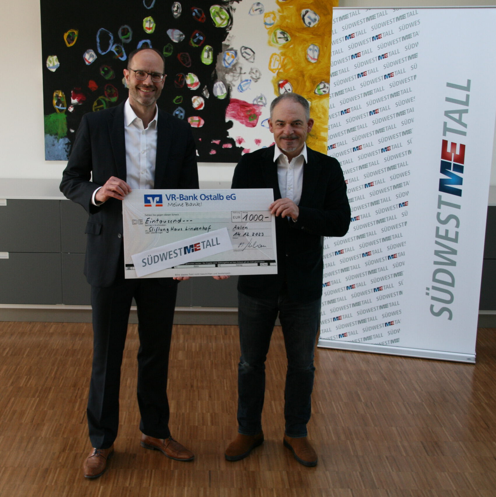 Zwei Männer in Anzügen halten stolz einen großen Spende-Scheck über 1.000 Euro von der VR-Bank Ostalb eG in einer Büroumgebung. Ein „Südwestmetall“-Banner und lebendige Bilder zieren den Hintergrund und fangen den Geist von Teilhabe und Gemeinschaftsunterstützung ein.