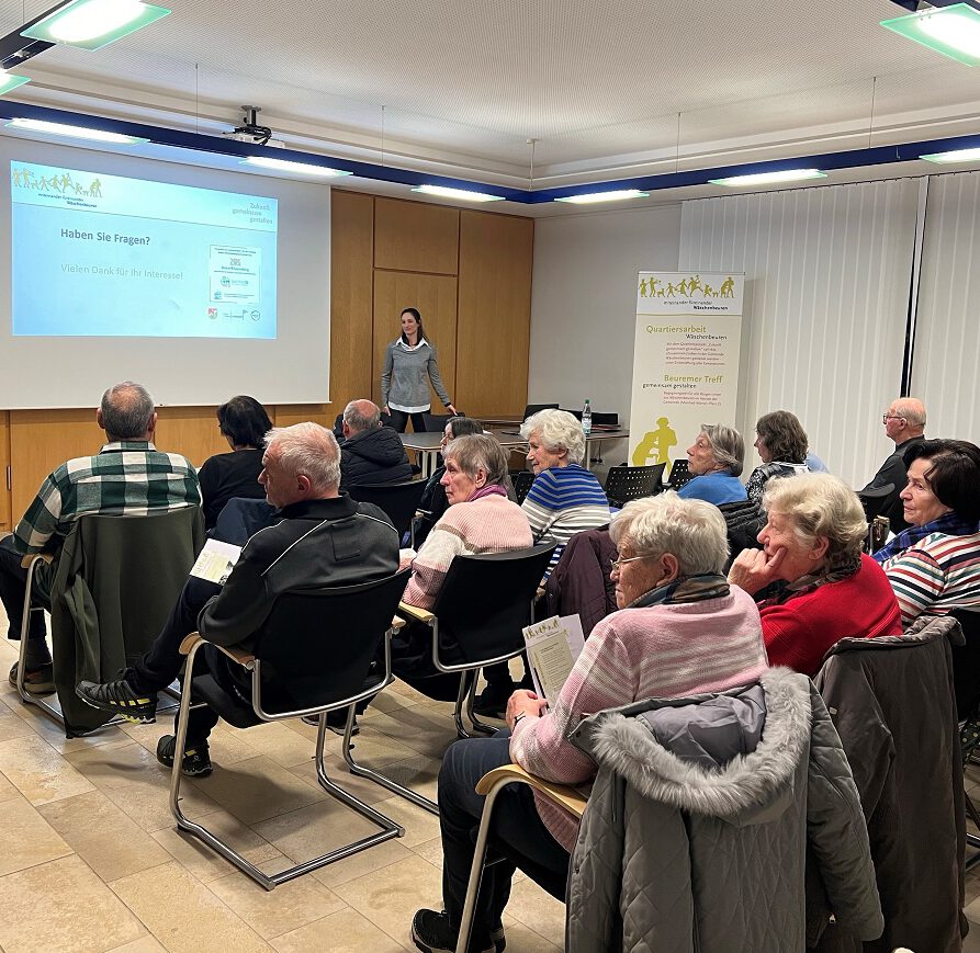In Wäschenbeuren feierte eine Gruppe älterer Teilnehmer im Rahmen des Quartiersprojekts „Ein Jahr“, indem sie während der Präsentation in einem Raum vor einer Projektionsleinwand saßen. Eine Frau steht vorne und spricht, während sie Broschüren halten. Im Hintergrund sind Banner mit Text und Grafiken zu sehen.